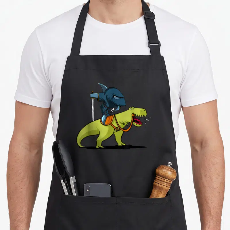 Shark dinosaur apron - Blue shark holding silver sword riding lime green dinosaur with orange saddle — shown on a black cotton canvas apron against a white background