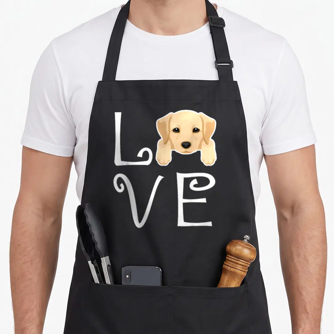 Owner retriever labrador apron - Cartoon beige puppy head and paws replacing the O in white LOVE lettering — shown on a black cotton canvas apron against a white background