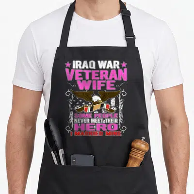 Iraq wife apron - Eagle holding ribbon surrounded by pink and white stacked text and stars — shown on a black cotton canvas apron against a white background