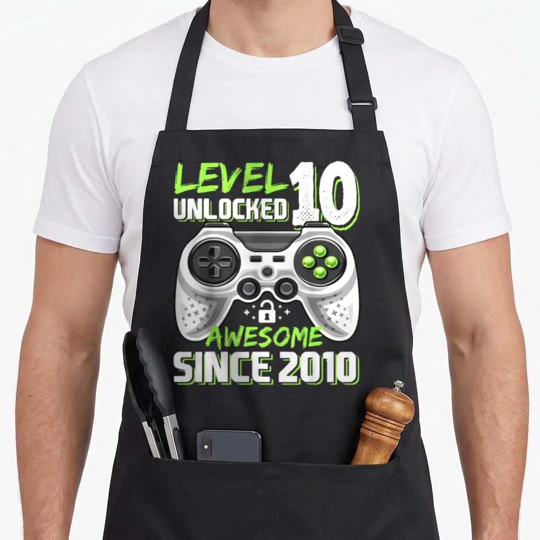 Unlocked video game apron - LEVEL 10 UNLOCKED game controller cartoon with green white grey black text and lock icon — shown on a black cotton canvas apron against a white background