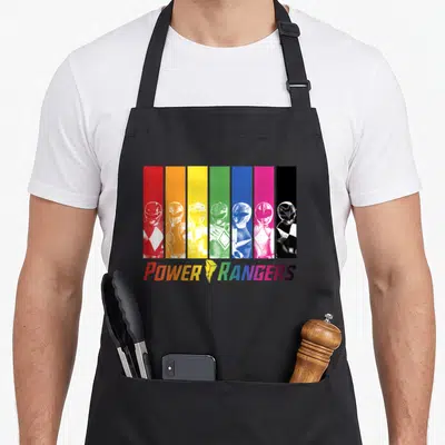 Character rainbow apron - Six Power Rangers in front of vertical red orange yellow green blue pink and black stripes — shown on a black cotton canvas apron against a white background