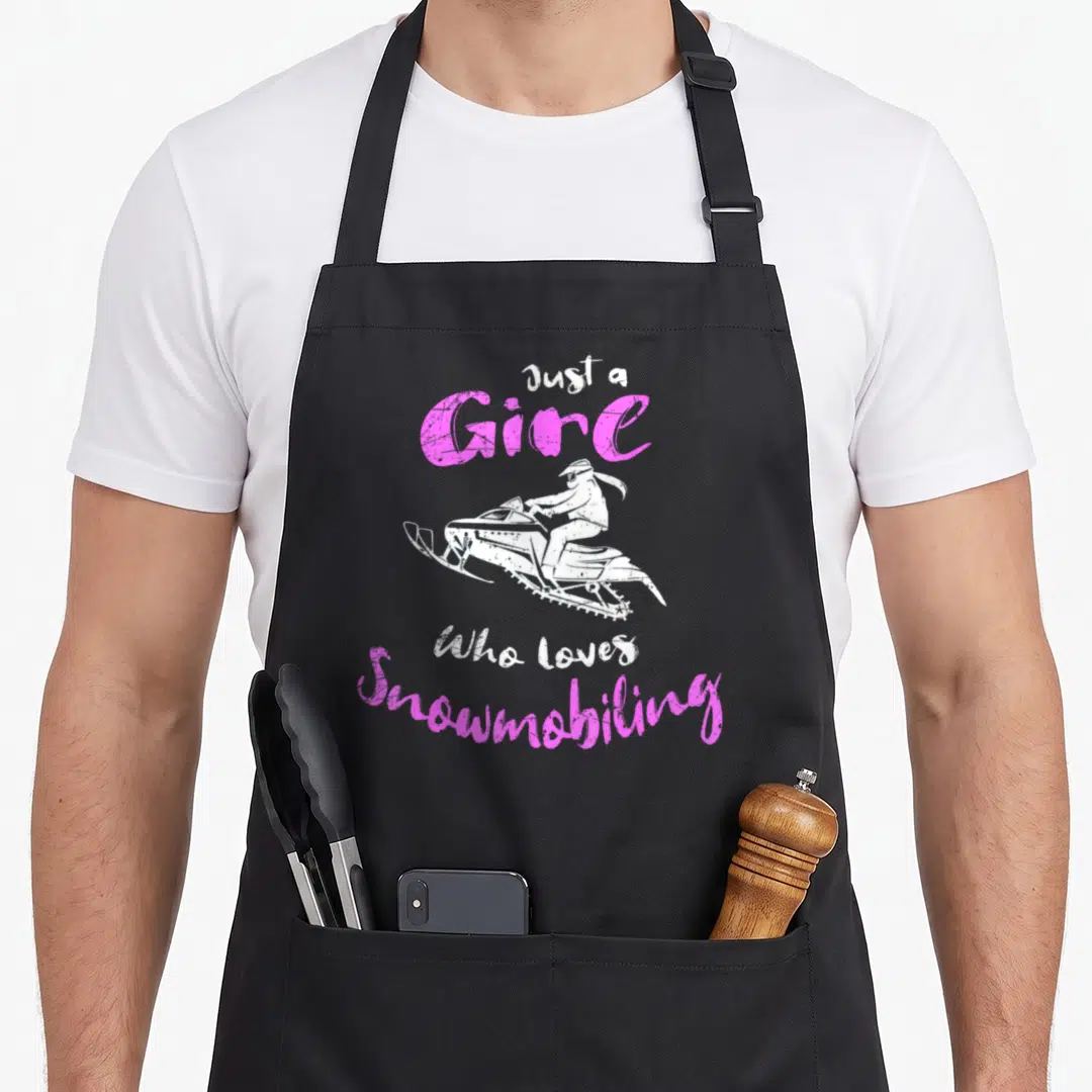 Costume snowmobile girl apron - Distressed pink text Just a Girl Who loves Snowmobiling above and below a white — shown on a black cotton canvas apron against a white background