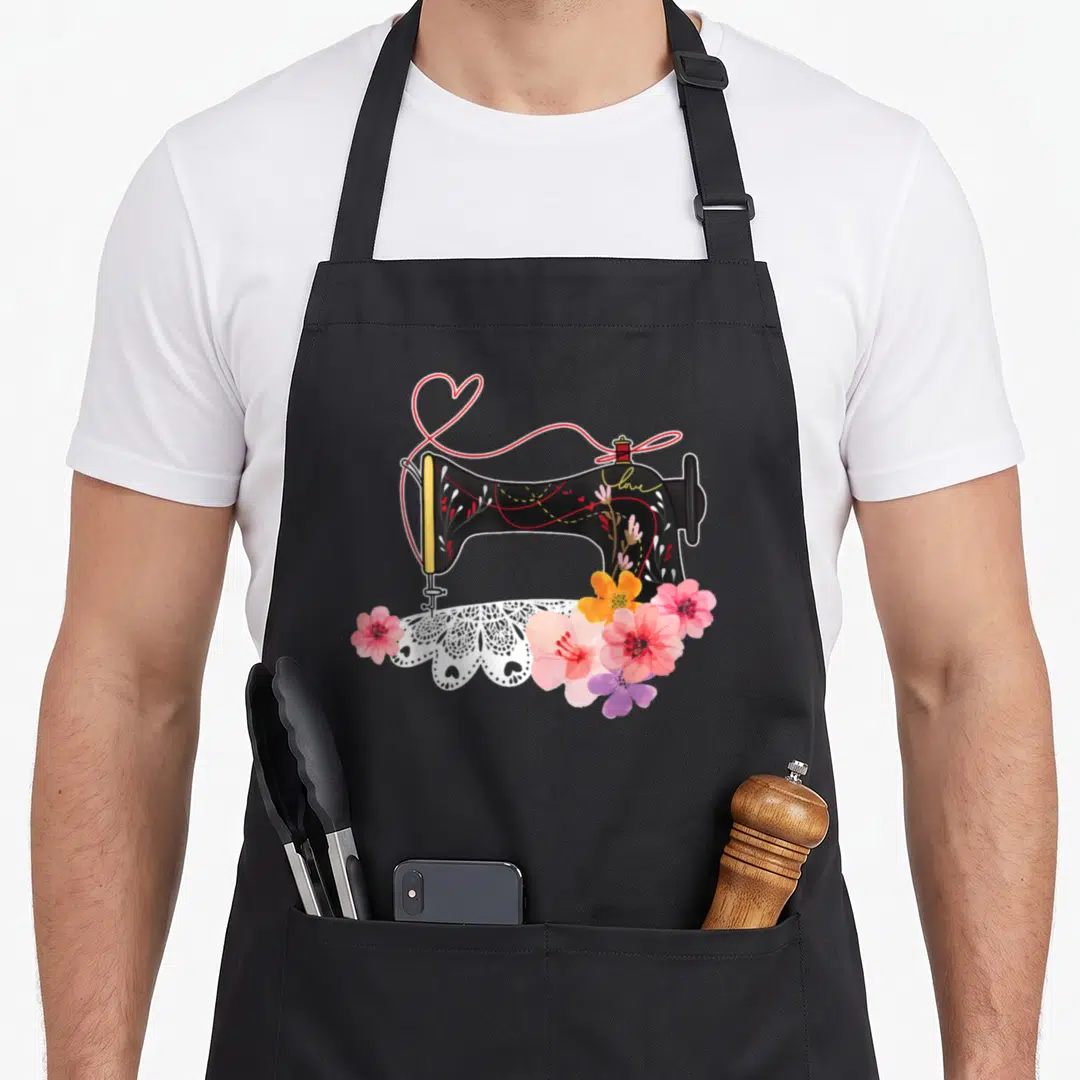 Sewing tailor apron - Black sewing machine with red thread heart, lace, and pink flowers printed with love text — shown on a black cotton canvas apron against a white background