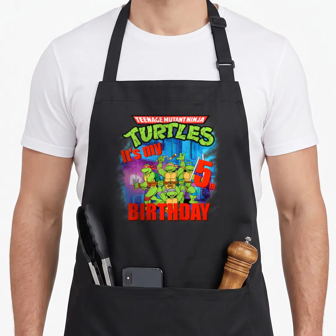 Fifth birthday apron - TMNT logo above four turtles holding weapons, red 'It's my 5th BIRTHDAY' text, green background — shown on a black cotton canvas apron against a white background