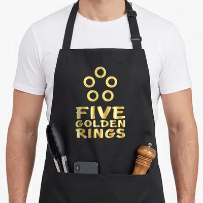 Ring christmas apron - Five golden rings text and five circular icons in gold typography — shown on a black cotton canvas apron against a white background