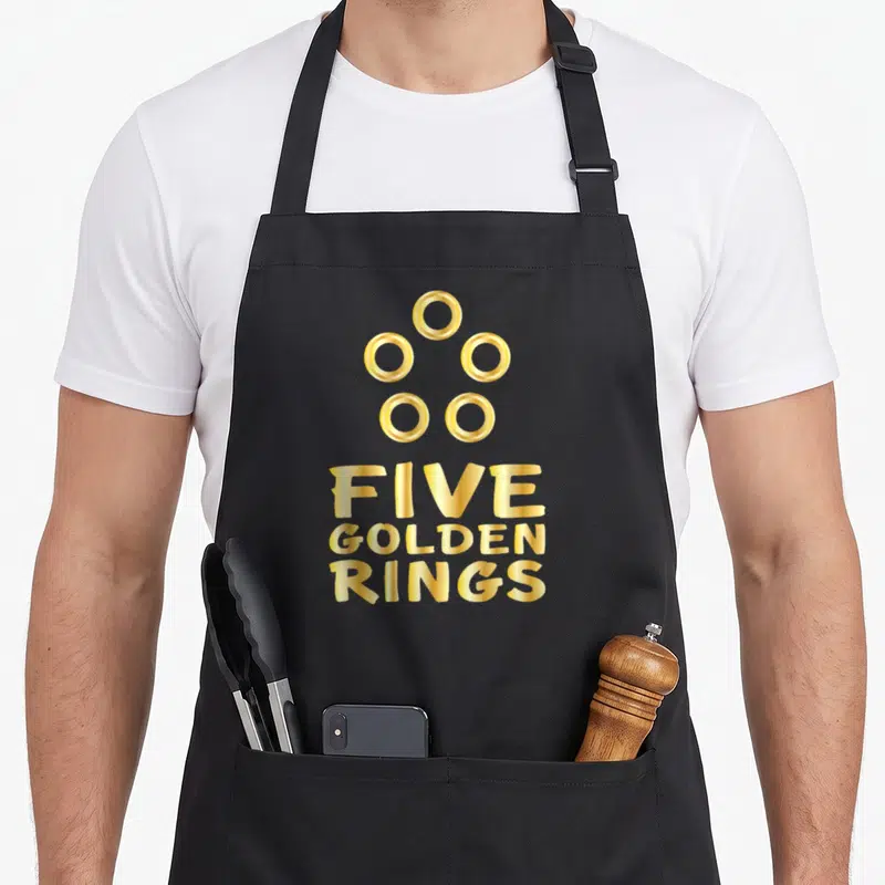 Ring christmas apron - Five golden rings text and five circular icons in gold typography — shown on a black cotton canvas apron against a white background