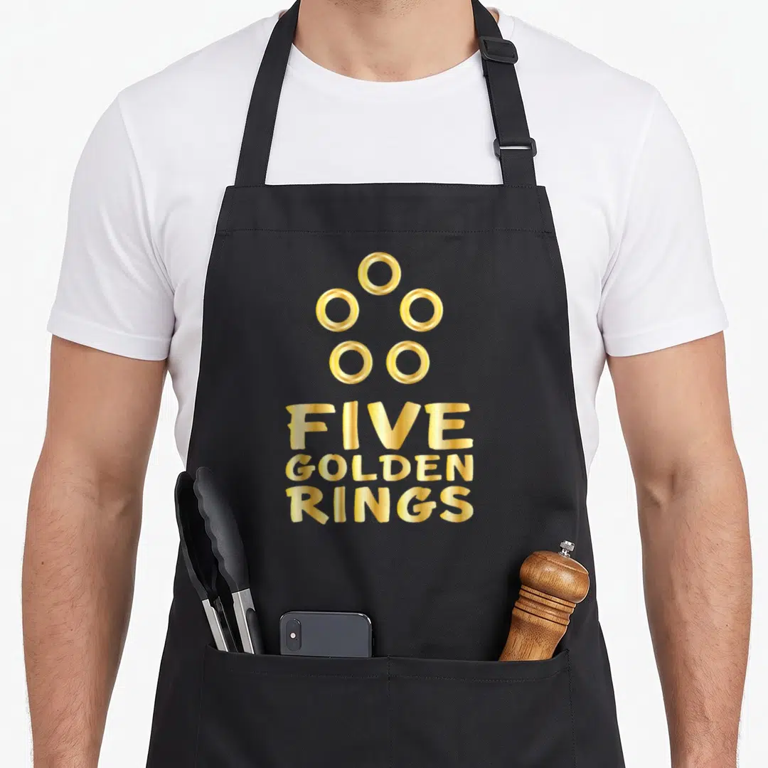 Ring christmas apron - Five golden rings text and five circular icons in gold typography — shown on a black cotton canvas apron against a white background