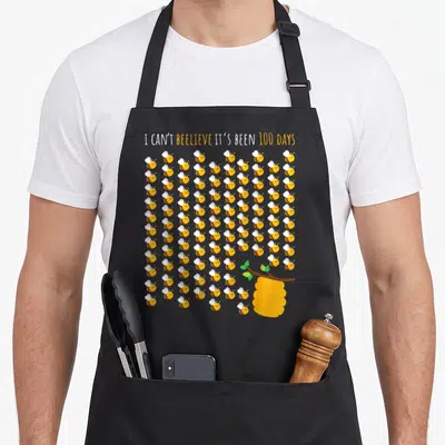 Tree bee aprons - 100 cartoon bees and beehive with 100 days school pun text — shown on a black cotton canvas apron against a white background