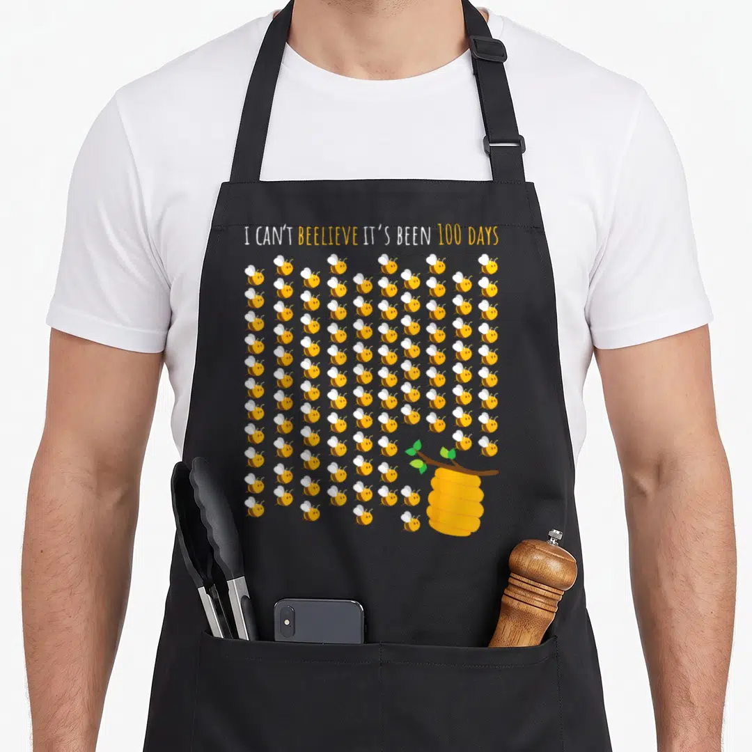 Tree bee aprons - 100 cartoon bees and beehive with 100 days school pun text — shown on a black cotton canvas apron against a white background