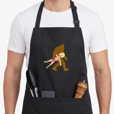 Blow bigfoot apron - Cartoon brown bigfoot carrying blonde woman in pink bikini — shown on a black cotton canvas apron against a white background