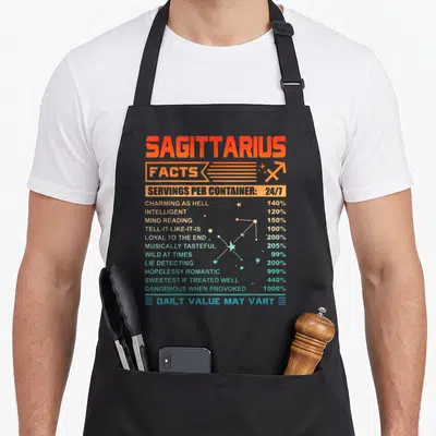 Sagittarius fact birthday apron - Orange and blue nutrition facts label style print listing Sagittarius traits — shown on a black cotton canvas apron against a white background