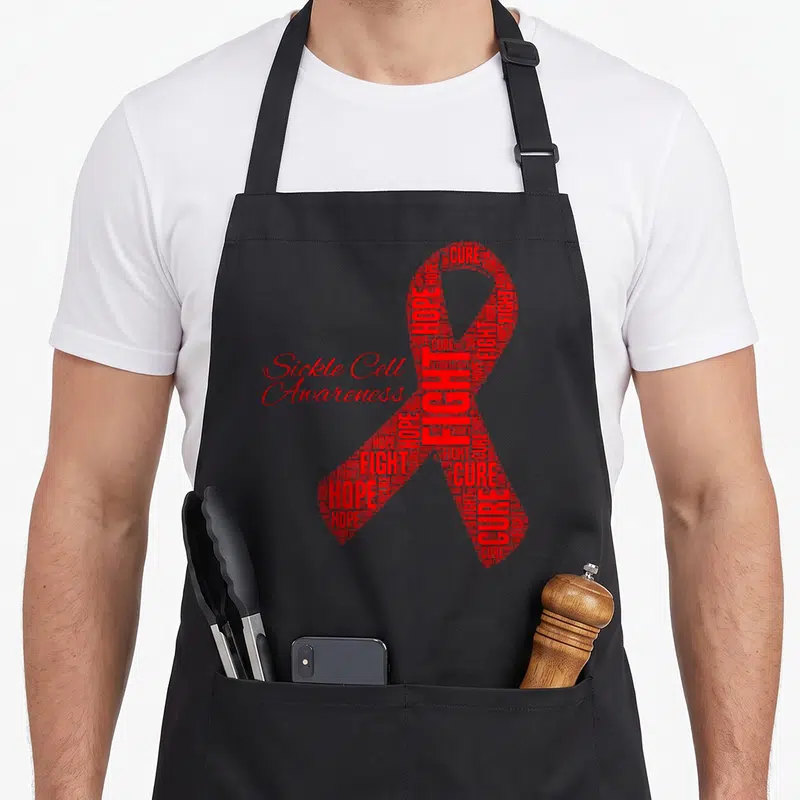Sickle ribbon apron - Red awareness ribbon formed by hope fight cure word cloud with Sickle Cell Awareness text — shown on a black cotton canvas apron against a white background