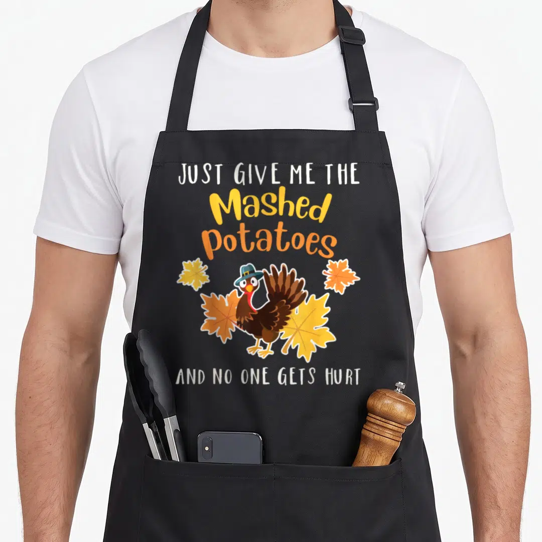 Give funny mashed apron - Cartoon turkey in blue hat with autumn leaves and text Just Give Me The Mashed Potatoes — shown on a black cotton canvas apron against a white background
