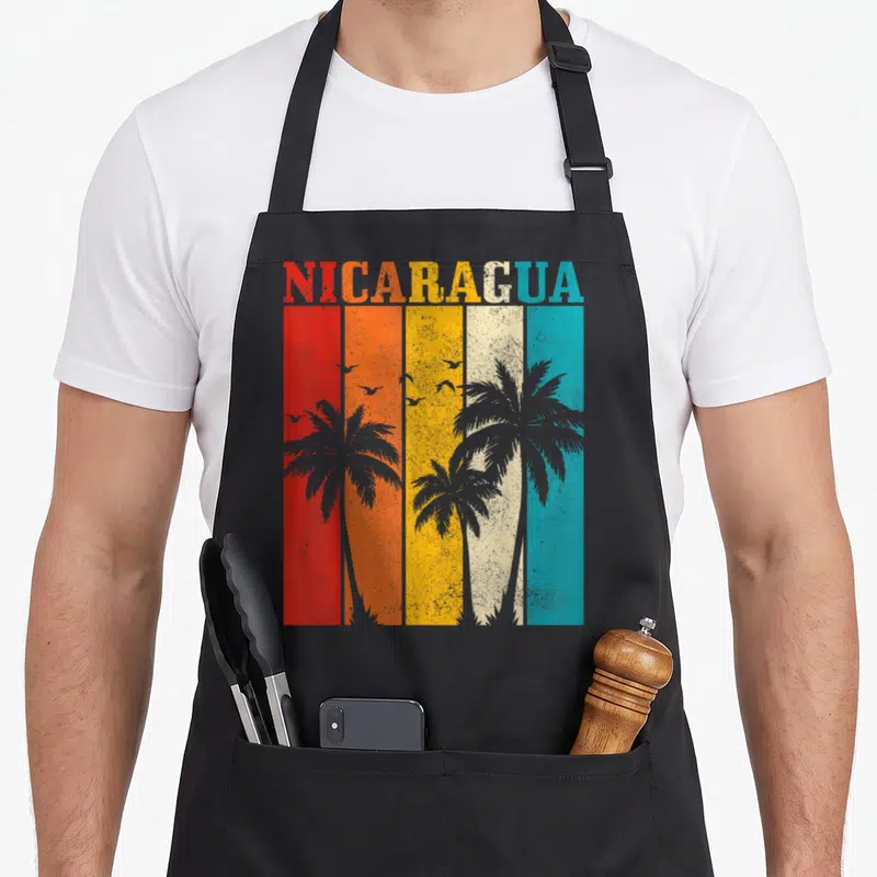 Nicaragua tree apron - NICARAGUA text over red orange yellow white blue stripes with grey palm trees and birds — shown on a black cotton canvas apron against a white background