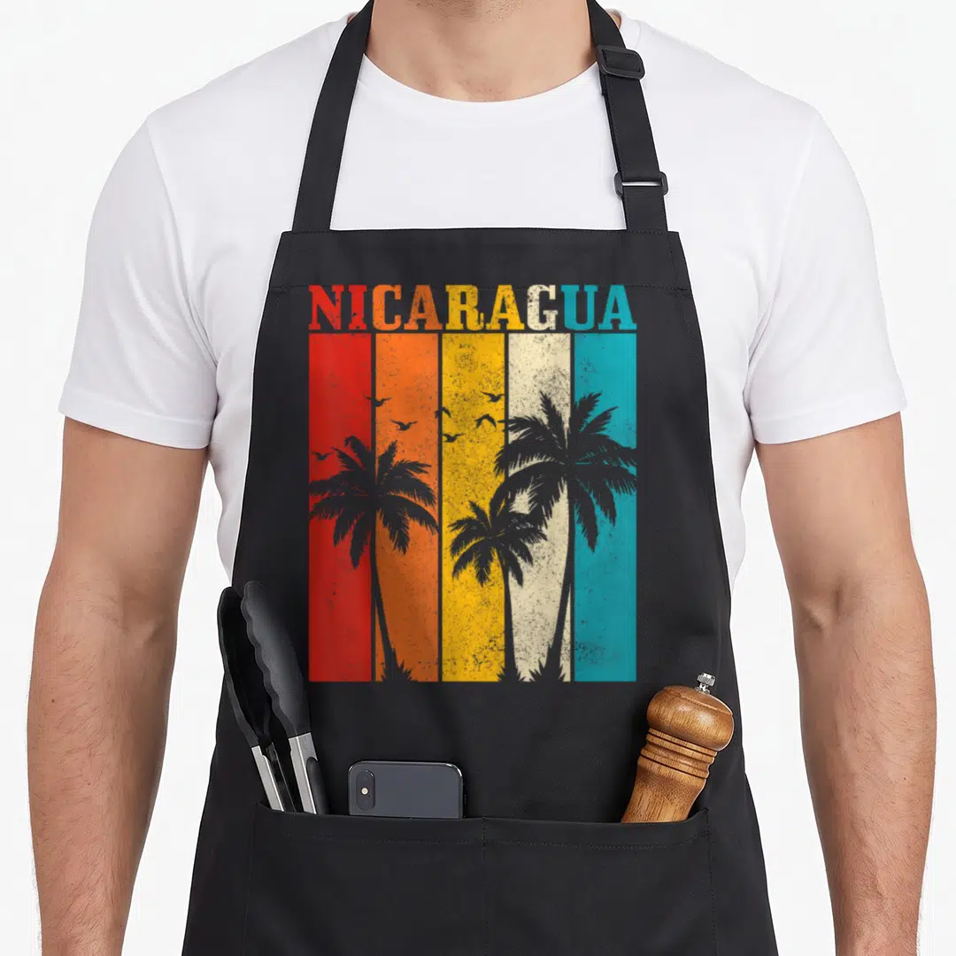 Nicaragua tree apron - NICARAGUA text over red orange yellow white blue stripes with grey palm trees and birds — shown on a black cotton canvas apron against a white background