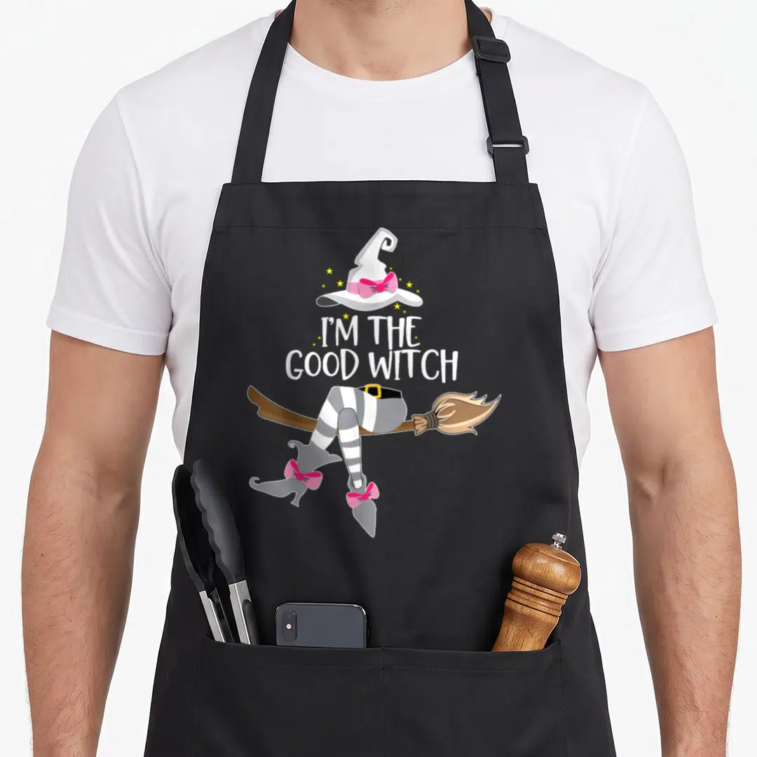 Funny costume halloween witch apron - Cartoon witch hat, I'M THE GOOD WITCH text, striped legs on broomstick with pink — shown on a black cotton canvas apron against a white background