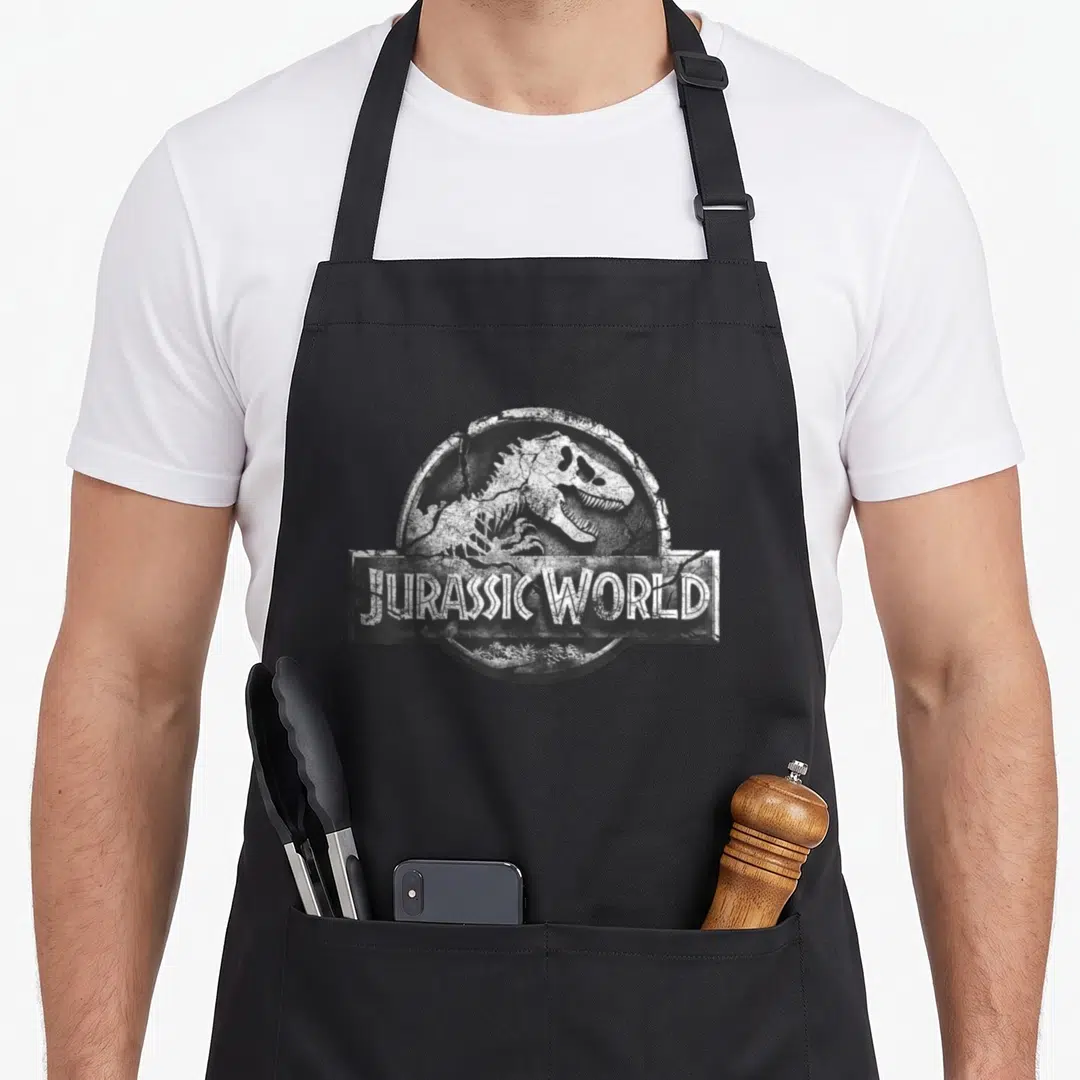 Jurassic stone apron - Distressed white dinosaur skeleton inside cracked stone circle with JURASSIC WORLD text bar — shown on a black cotton canvas apron against a white background