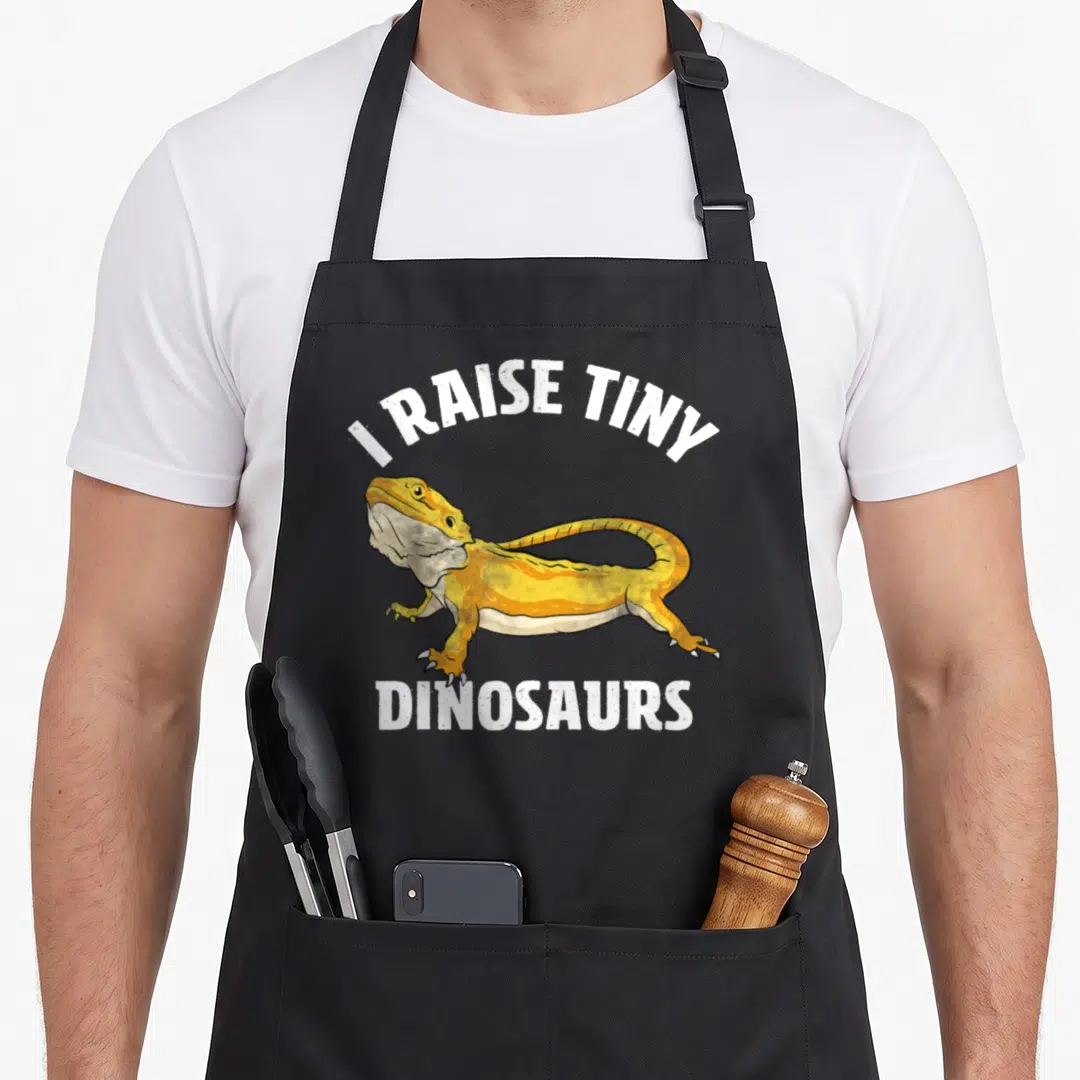 Beard tiny chef apron - Yellow bearded dragon with I Raise Tiny Dinosaurs text in white cartoon font — shown on a black cotton canvas apron against a white background