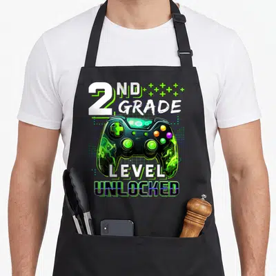 Gamer 2nd apron - 2nd Grade Level Unlocked text above green and black game controller on grid background — shown on a black cotton canvas apron against a white background