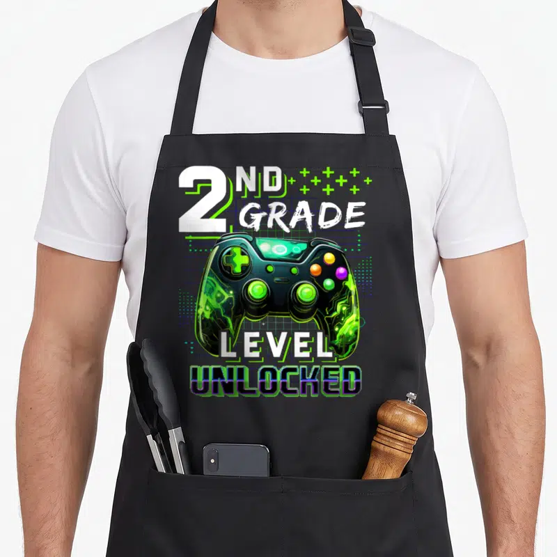 Gamer 2nd apron - 2nd Grade Level Unlocked text above green and black game controller on grid background — shown on a black cotton canvas apron against a white background