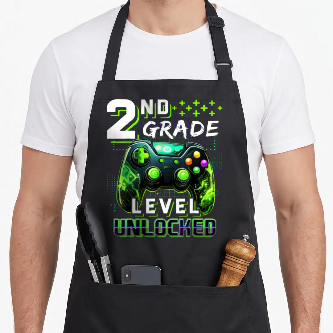Gamer 2nd apron - 2nd Grade Level Unlocked text above green and black game controller on grid background — shown on a black cotton canvas apron against a white background