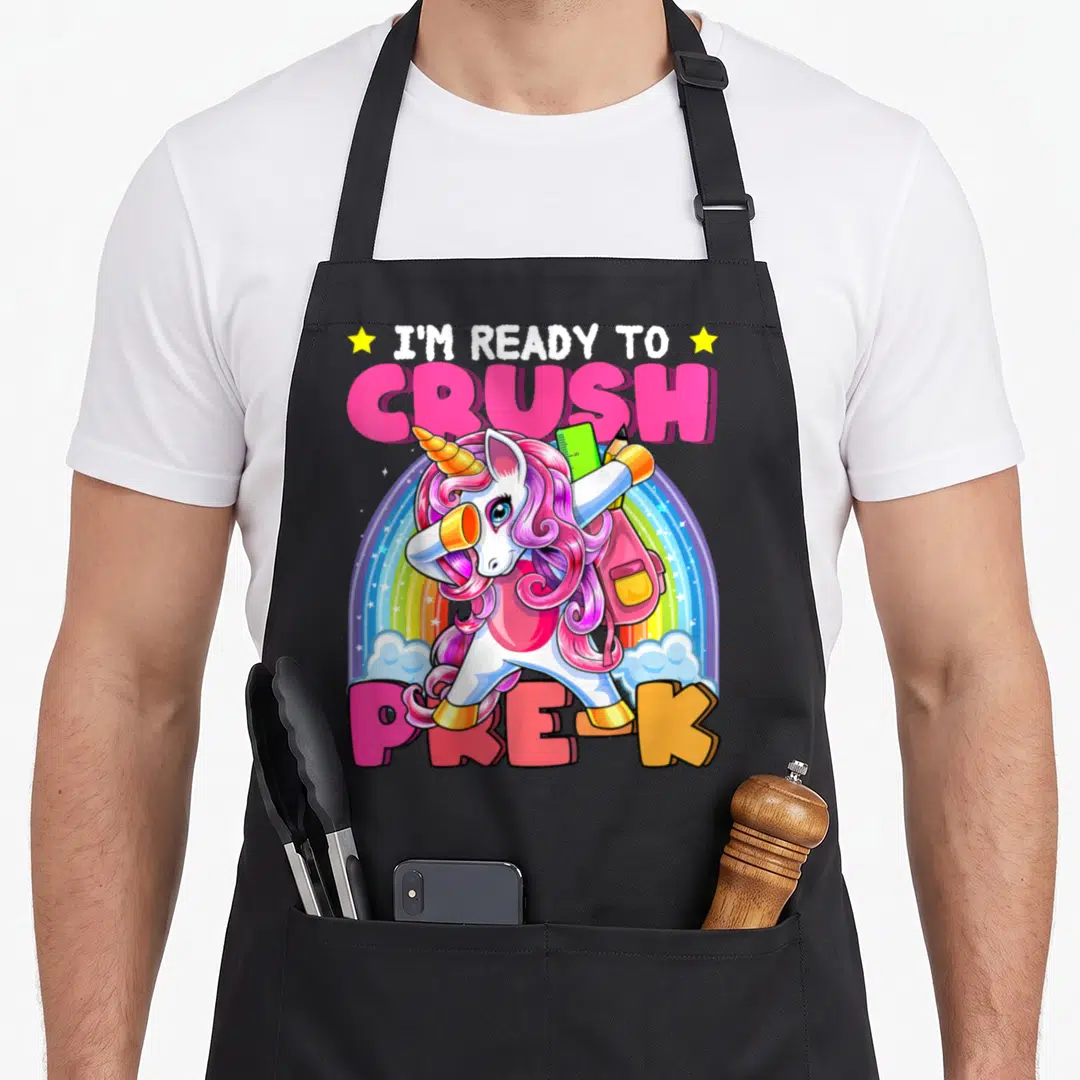 Crush pre unicorn apron - Dabbing unicorn with backpack and ruler in front of rainbow with I'M READY TO CRUSH PRE-K text — shown on a black cotton canvas apron against a white background