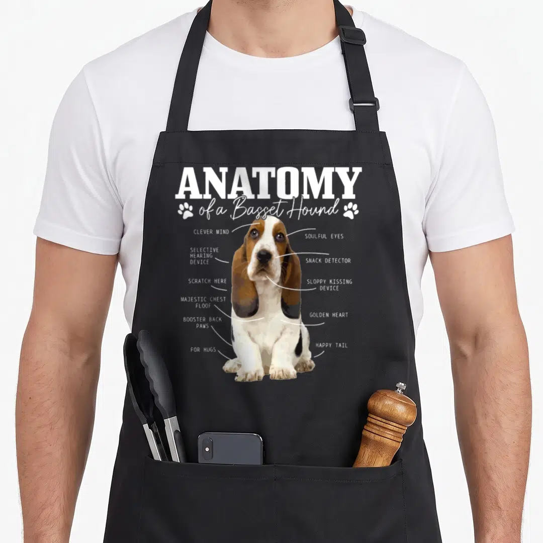Anatomy basset dog apron - Diagram of a basset hound with labels for snack detector and sloppy kissing device — shown on a black cotton canvas apron against a white background
