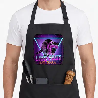 Waschbr raccoon apron - Raccoon in pink sunglasses inside neon blue triangle with LIVE FAST EAT TRASH text on purple — shown on a black cotton canvas apron against a white background
