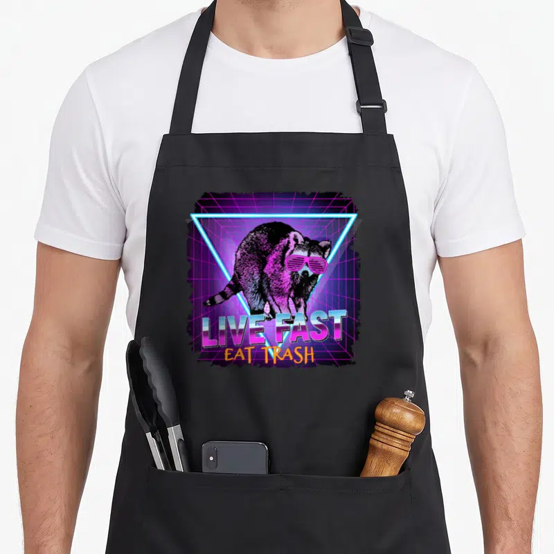 Waschbr raccoon apron - Raccoon in pink sunglasses inside neon blue triangle with LIVE FAST EAT TRASH text on purple — shown on a black cotton canvas apron against a white background