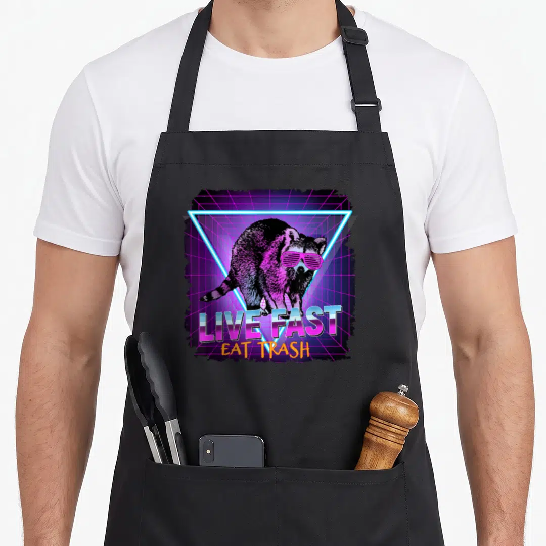 Waschbr raccoon apron - Raccoon in pink sunglasses inside neon blue triangle with LIVE FAST EAT TRASH text on purple — shown on a black cotton canvas apron against a white background