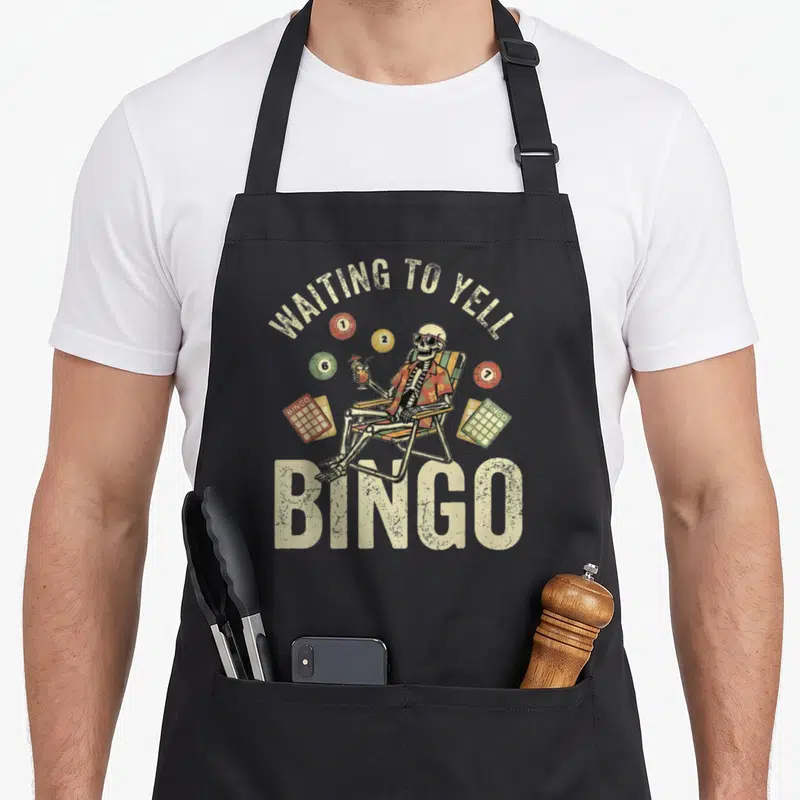 Bingo skeleton apron - Cartoon skeleton in beach chair with colorful bingo balls and cards under arched text — shown on a black cotton canvas apron against a white background