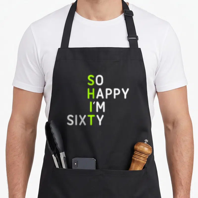 Sixty 60th birthday apron - Stacked white text with green highlights spelling SHIT vertically on So Happy I'm Sixty — shown on a black cotton canvas apron against a white background