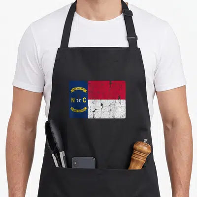 Souvenir carolina apron - Distressed NC flag with blue hoist, red white stripes, and yellow date banners — shown on a black cotton canvas apron against a white background