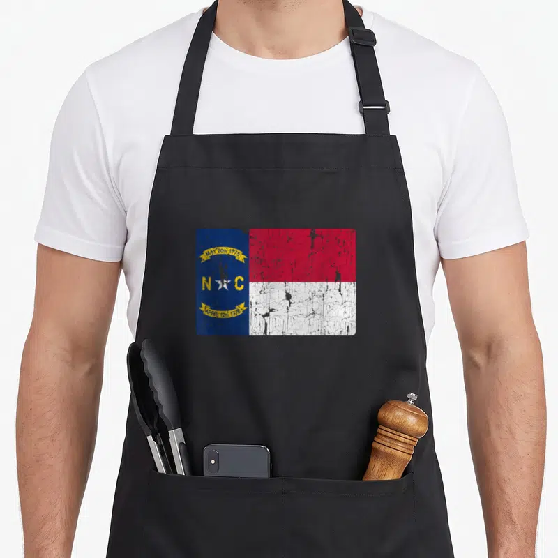 Souvenir carolina apron - Distressed NC flag with blue hoist, red white stripes, and yellow date banners — shown on a black cotton canvas apron against a white background