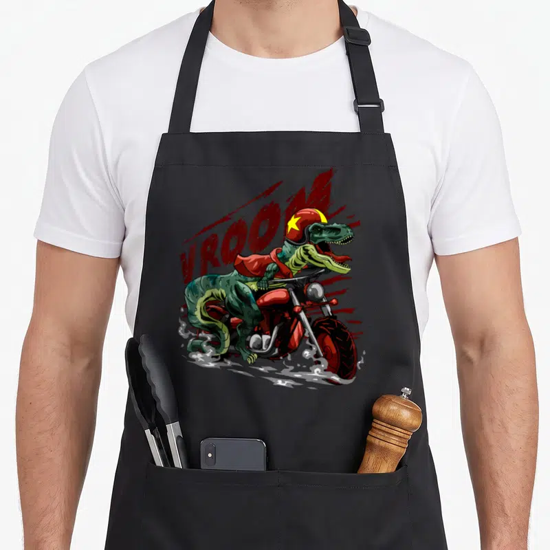 Motorcycle dinosaur apron - Tyrannosaurus Rex riding red motorcycle wearing helmet with VROOM text in red and green — shown on a black cotton canvas apron against a white background