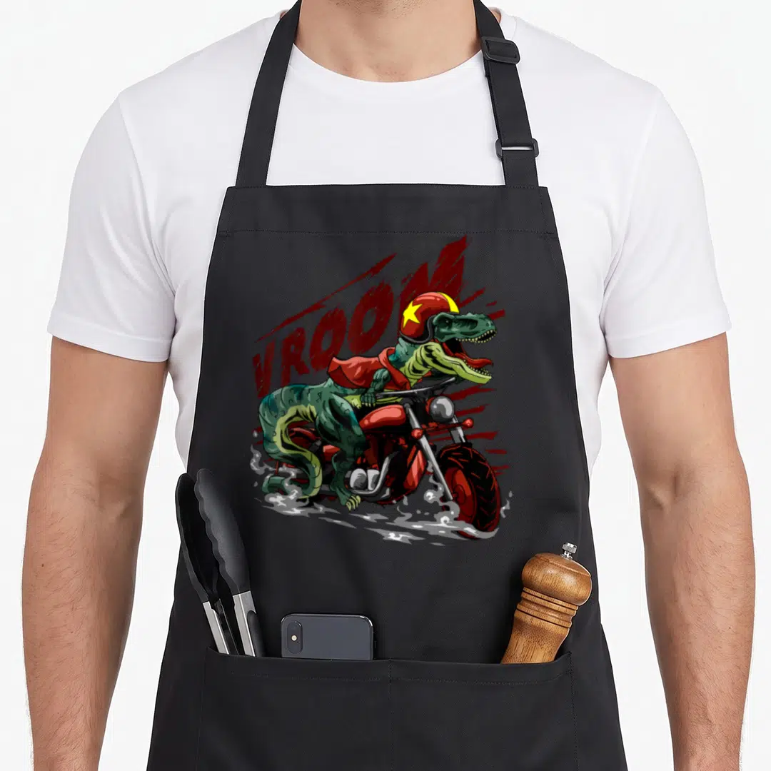Motorcycle dinosaur apron - Tyrannosaurus Rex riding red motorcycle wearing helmet with VROOM text in red and green — shown on a black cotton canvas apron against a white background