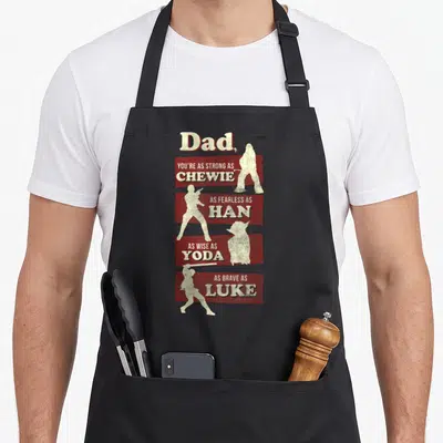 Strong father day apron - Dad text compared to Chewie Han Yoda and Luke with alternating cream silhouettes on red — shown on a black cotton canvas apron against a white background