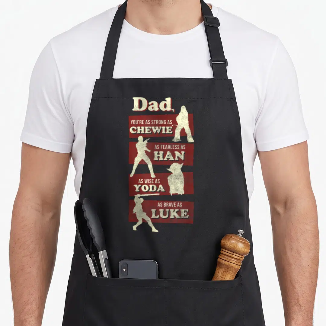 Strong father day apron - Dad text compared to Chewie Han Yoda and Luke with alternating cream silhouettes on red — shown on a black cotton canvas apron against a white background