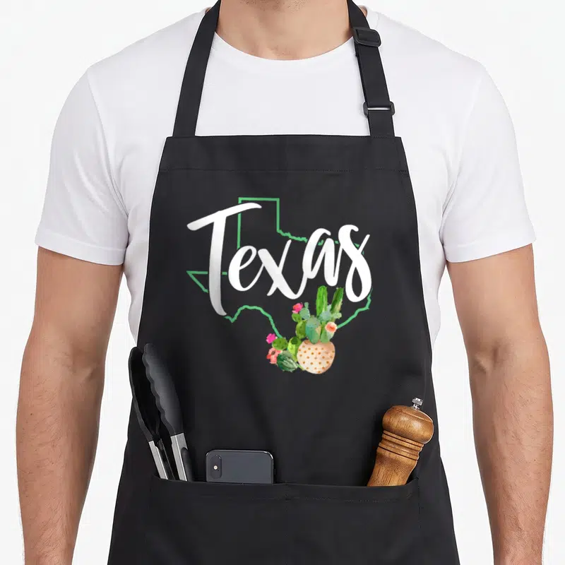 Pride cactus apron - Texas script over green state outline with potted cactus and pink flowers in watercolor style — shown on a black cotton canvas apron against a white background