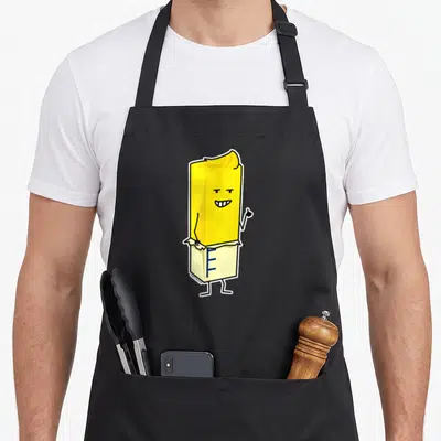 Buttered butter apron - Yellow cartoon butter stick with face and thumbs up inside white wrapper with blue markings — shown on a black cotton canvas apron against a white background