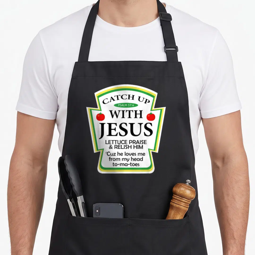 Ketchup catch up with jesus apron - Ketchup label graphic with CATCH UP WITH JESUS text and two red tomatoes flanking — shown on a black cotton canvas apron against a white background