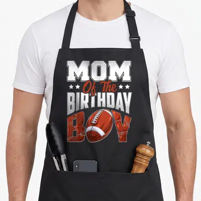 Football family aprons - Stacked text Mom Of The Birthday Boy with red football replacing O and white stars — shown on a black cotton canvas apron against a white background