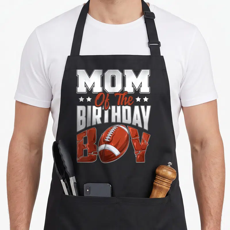 Football family aprons - Stacked text Mom Of The Birthday Boy with red football replacing O and white stars — shown on a black cotton canvas apron against a white background