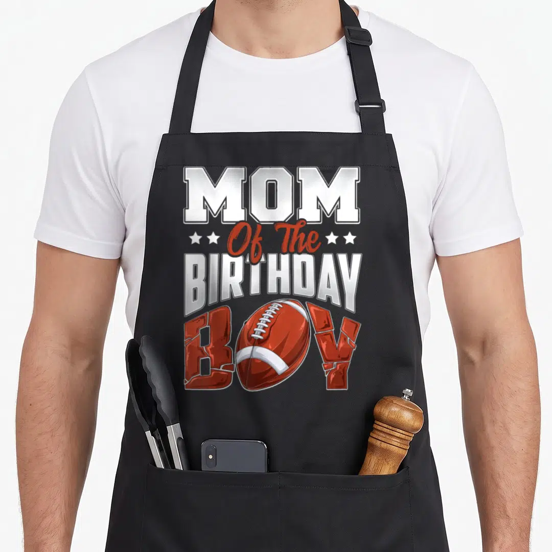 Football family aprons - Stacked text Mom Of The Birthday Boy with red football replacing O and white stars — shown on a black cotton canvas apron against a white background