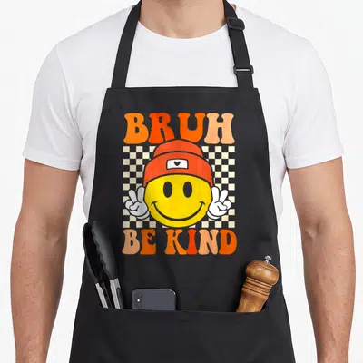 Bruh unity day apron - Smiley face in orange beanie with peace signs and Bruh Be Kind text on checkerboard — shown on a black cotton canvas apron against a white background
