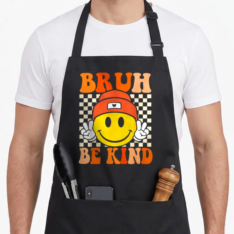Bruh unity day apron - Smiley face in orange beanie with peace signs and Bruh Be Kind text on checkerboard — shown on a black cotton canvas apron against a white background