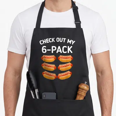 Check hot dog apron - CHECK OUT MY 6-PACK text above six cartoon hot dogs in red and yellow — shown on a black cotton canvas apron against a white background