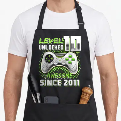 10th video game apron - Cartoon game controller with LEVEL 10 UNLOCKED AWESOME SINCE 2011 text and lime green dots — shown on a black cotton canvas apron against a white background