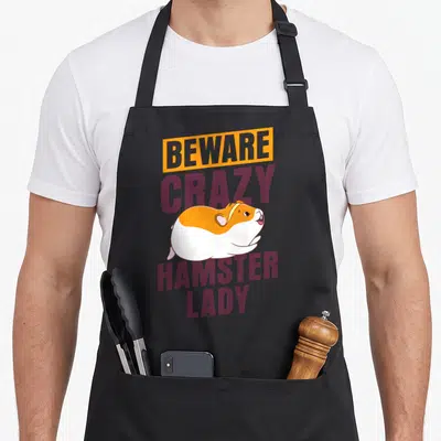 Vintage beware crazy hamster lady apron - Stacked text BEWARE CRAZY HAMSTER LADY with cartoon hamster in orange — shown on a black cotton canvas apron against a white background