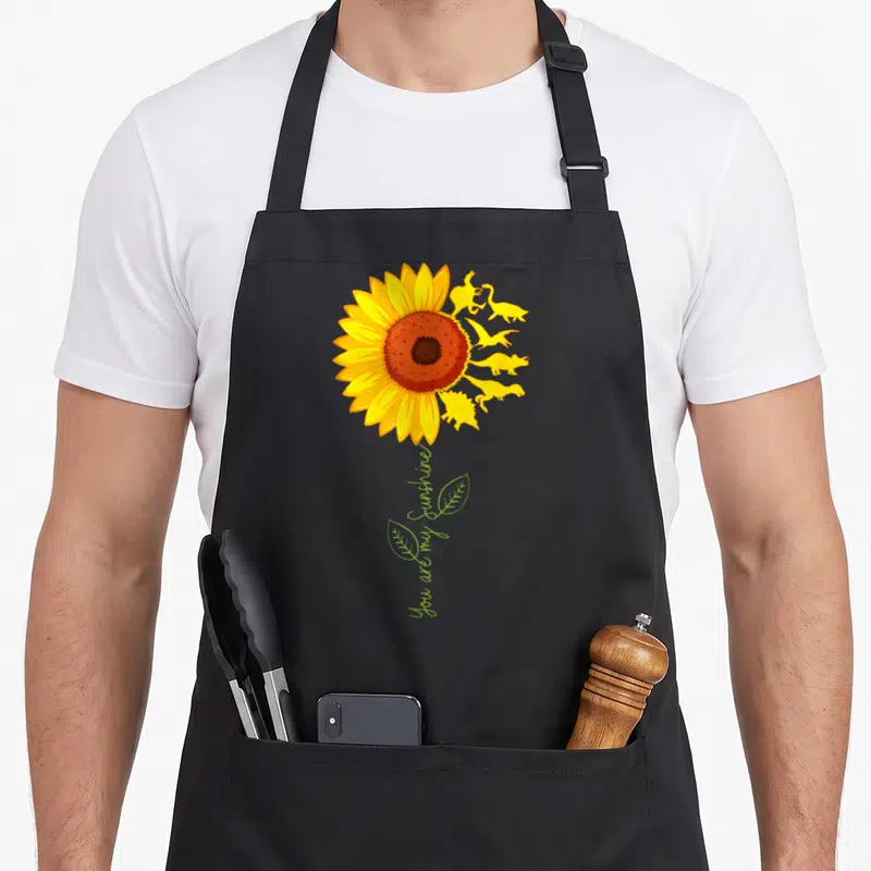 Bist sunflower apron - You are my sunshine text below yellow sunflower and dinosaur silhouettes — shown on a black cotton canvas apron against a white background